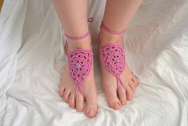 Nahaufnahme von Füßen mit rosa gehäkelten Barfußsandalen mit Perlen auf weißer Decke.