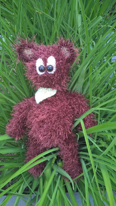 Flauschiger gehäkelter Teddybär in rötlich-braunem Effektgarn mit weißer Brustapplikation, liegend im Gras