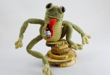 Hellgrüner gehäkelter Amigurumi-Frosch mit roter Zunge und Fliege auf Holzscheiben vor weißem Hintergrund