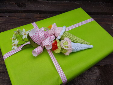 Grün verpacktes Geschenk mit rosa-weiß kariertem Band, zwei gehäkelten Amigurumi-Schultüten und weißen Blüten auf dunklem Holzhintergrund.