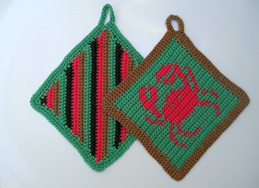 Zwei gehäkelte quadratische Topflappen in Grün, Rot und Braun; einer mit rotem Krebsmotiv, einer gestreift