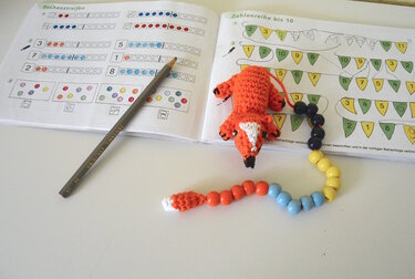 Gehäkelter orange-weißer Fuchs mit bunter Perlenkette auf geöffnetem Rechenheft neben einem Bleistift.