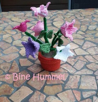 Gehäkelter Blumentopf mit rosa, pinken, lila und weißen Blumen auf einem steinernen Tisch