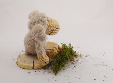 Gehäkeltes Amigurumi-Schäfchen aus flauschigem Garn sitzt auf Holzscheiben neben Moos