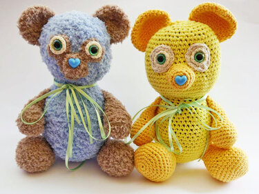 Zwei gehäkelte Amigurumi-Teddybären, flauschig blau und gelb, mit grünen Bändern und blauer Herznase.