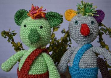 Zwei gehäkelte Amigurumi-Figuren, grün und grau, mit bunten Nasen und Haarbüscheln