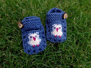 Zwei dunkelblaue, gestrickte Babyschuhe mit Eulenmotiv, aufgenähten Wackelaugen und Holzknöpfen auf Gras