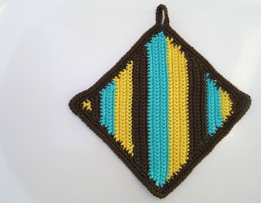 Gehäkelter Topflappen in Rautenform mit dunkelbraunem Rand und gelben sowie türkisen Streifen auf hellem Hintergrund.