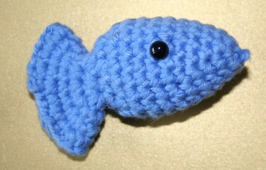 Blauer gehäkelter Amigurumi-Fisch mit schwarzem Knopfauge auf hellem Hintergrund.