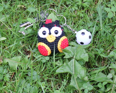 Gehäkelte Eule (schwarz, rot, gelb) als Schlüsselanhänger mit kleinem Fußball-Anhänger auf Gras neben Schlüsseln.