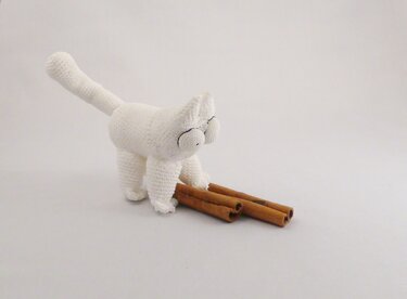 Weißer gehäkelter Katzen-Amigurumi mit drei Zimtstangen auf hellem Untergrund