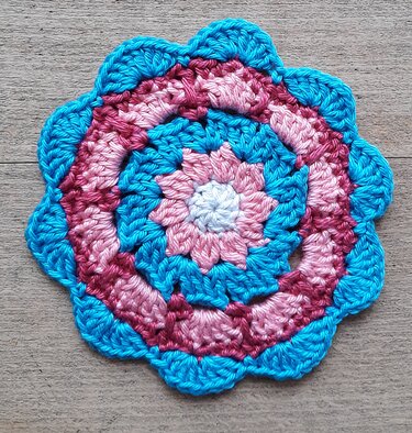 Runder gehäkelter Untersetzer in blau und rosa mit muschelförmigem Rand auf Holz