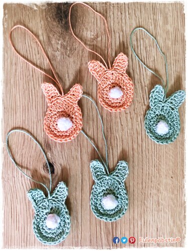 Fünf gehäkelte Hasenanhänger in Pastell (grün und koralle) mit weißen Bommeln auf Holzuntergrund