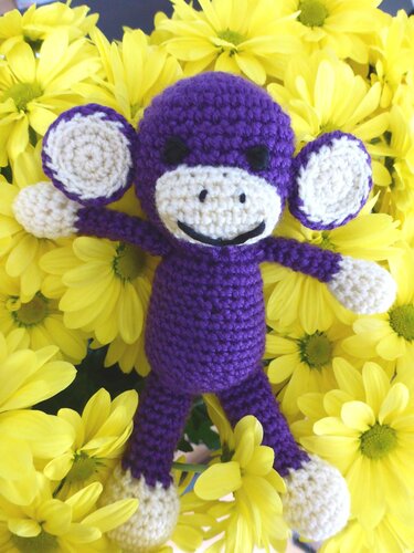 Gehäkelter lila Amigurumi-Affe mit cremefarbenem Gesicht und Gliedmaßen auf gelben Blumen