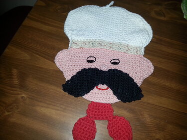 Crocheted chef potholder with white hat, pink face, black mustache, and red neck detail on a wooden table.