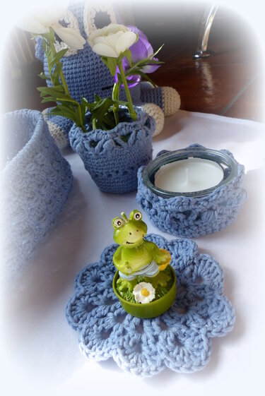 Gehäkelte hellblaue Deko auf einem Tisch: Blumentopfhülle mit Blumen, Teelichthalter mit Häkelmanschette, Untersetzer und Froschfigur.