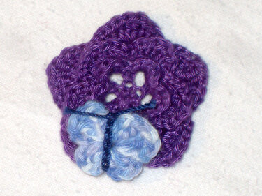 Gehäkelte lila Blume mit hellblauem Schmetterling.