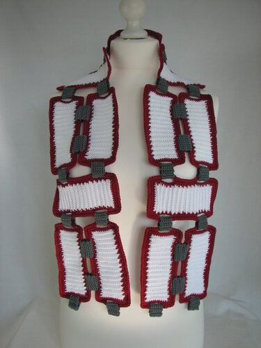 Gehäkelter Schal aus weißen rechteckigen Modulen mit roten Rändern und grauen Verbindungsstücken auf Schaufensterpuppe