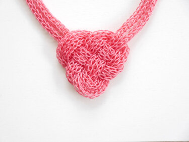 Kunstvoll gestricktes, rosafarbenes Herz als zentrales Element einer Halskette auf weißem Hintergrund; Strickstruktur deutlich erkennbar.