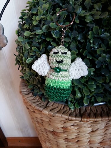 Kleines grünes Amigurumi‑Engelchen mit weißen Flügeln an Schlüsselring vor Pflanzenhintergrund