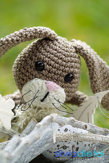 Brauner Amigurumi-Hase mit langen Schlappohren, heller Schnauze und rosa Nase zwischen Zweigen und gesprenkeltem Deko-Ei.