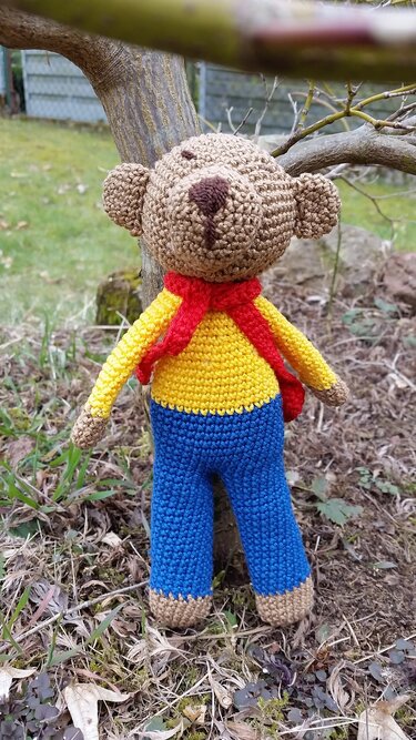 Gehäkelter brauner Teddybär mit gelbem Shirt, blauer Hose und rotem Schal
