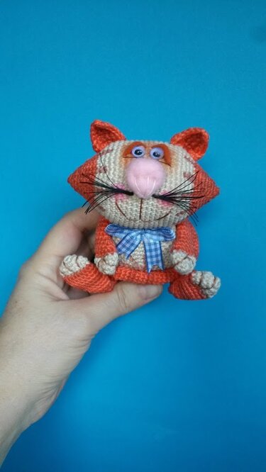 Hand hält gehäkelte Amigurumi-Katze in Orange und Creme mit rosa Nase und blau kariertem Schleifchen vor türkisfarbenem Hintergrund