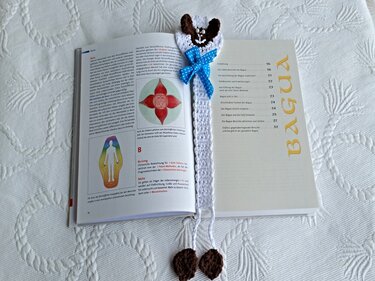 Weißes gehäkeltes Lesezeichen mit braun-weißem Schafkopf, blauer gepunkteter Schleife und zwei gehäkelten Endstücken in einem geöffneten Buch