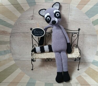 Gehäkelter Amigurumi‑Waschbär in Grau und Schwarz auf einer Miniaturbank, hält ein Schild mit der Aufschrift „Rocco“.