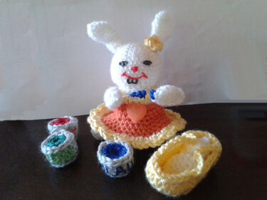 Weißes gehäkeltes Amigurumi-Häschen mit orange-gelbem Kleid, Herz-Applikation, Blume am Ohr und kleinen gehäkelten Accessoires