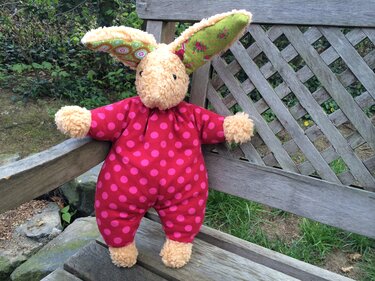 Handgenähter Plüschhase im pinken Punkte-Overall mit gemusterten Ohren auf einer Holzbank im Freien