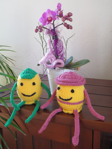 Zwei gelbe gestrickte Ostereier mit lächelnden Gesichtern auf einem Holzsideboard vor einer violetten Orchidee
