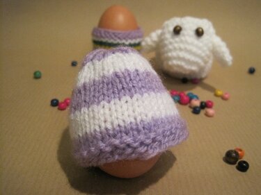 Zwei Eier mit gestrickten Eierwärmern (lila-weiß gestreift), kleine weiße Strickfigur und bunte Perlen