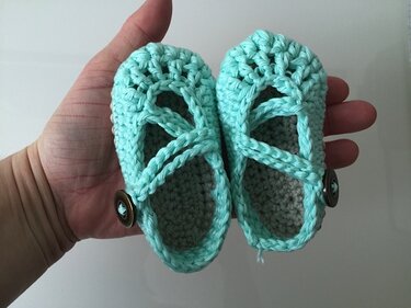Zwei mintgrüne gehäkelte Baby-Ballerinas mit Knöpfen und grauen Innensohlen in einer Hand.