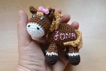 Detailaufnahme eines kleinen braun-weißen Amigurumi-Pferdes mit rosa Schleife und gesticktem Namen Fiona