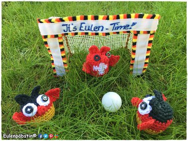 Drei gehäkelte Eulen in Schwarz, Rot und Gold mit Ball und Mini-Tor auf Rasen, Banner ‚It's Eulen - Time!‘.