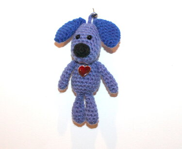Gehäkelter lila Amigurumi-Hund mit blauen Ohren, schwarzer Nase und rotem Herzanhänger an einer Schlaufe.