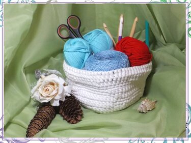 Weißer gehäkelter Korb mit bunten Wollknäueln, Häkelnadeln und Schere auf grünem Stoff.