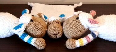 Drei gehäkelte Amigurumi-Schafe unterschiedlicher Größe mit weißen Körpern, beigebraunen Köpfen und bunten gestreiften Beinen auf dunklem Untergrund