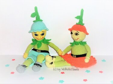 Zwei gehäkelte Amigurumi-Puppen in Pastellfarben halten Händchen; eine mit blauem Hut und Shorts, die andere mit korallfarbenem Rock