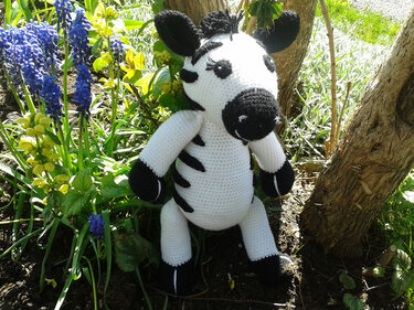 Handgehäkeltes schwarz-weißes Zebra-Amigurumi zwischen blauen Traubenhyazinthen neben einem Baumstamm