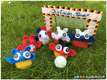 Sechs gehäkelte Eulenfiguren vor Mini-Fußballtor auf Gras mit weißem Ball