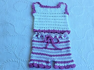 Zweiteiliges gehäkeltes Baby-Set in Weiß und Lila: ärmelloses Oberteil mit Knöpfen an den Schultern und gestreifte Shorts mit Kordelzug, auf hellblauem Untergrund.