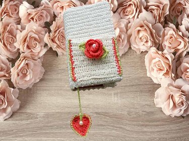 Cremefarbene gehäkelte Hülle mit roter Rose und Herzanhänger auf einem Holztisch, umgeben von rosafarbenen Rosen.