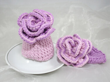 Zwei handgehäkelte Rosen in Lila und Rosa, eine als Eierwärmer auf einer Untertasse, die andere flach liegend mit Blatt.
