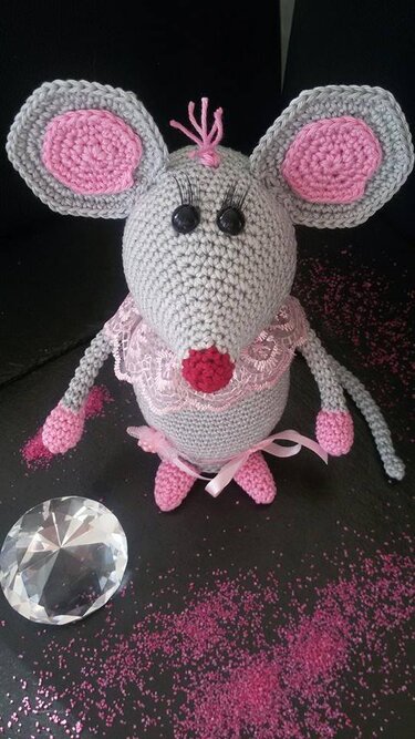 Gehäkelte graue Amigurumi-Maus mit rosa Innenohren, Spitzenkragen, Schleife und roter Nase auf dunklem, glitzerndem Untergrund mit Kristall.