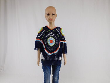 Schaufensterpuppe trägt einen dunkelblauen, gehäkelten Kinderponcho mit mehrfarbigem, kreisförmigem Motiv und Fransen über weißem Oberteil und Jeans.