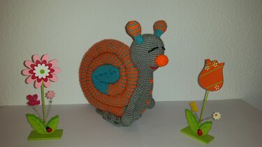 Gehäkelte Amigurumi-Schnecke mit gestreifter Spiral-Schale in Orange und Türkis neben zwei Holzblumen