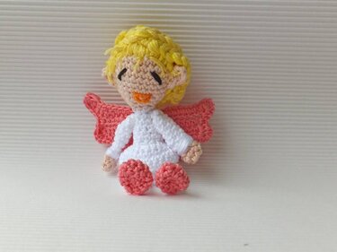 Gehäkeltes Amigurumi‑Engelchen mit gelbem Haar, weißem Kleid und rosafarbenen Flügeln, sitzend
