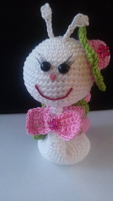 Weiße gehäkelte Amigurumi-Schnecke mit rosa Schleife, schwarzen Wimpern und herzförmiger Nase